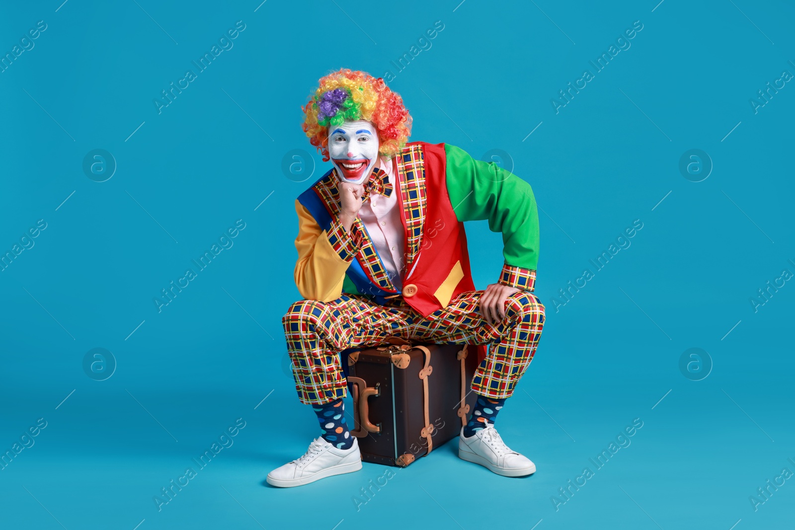 Photo of Happy clown sitting on suitcase against blue background