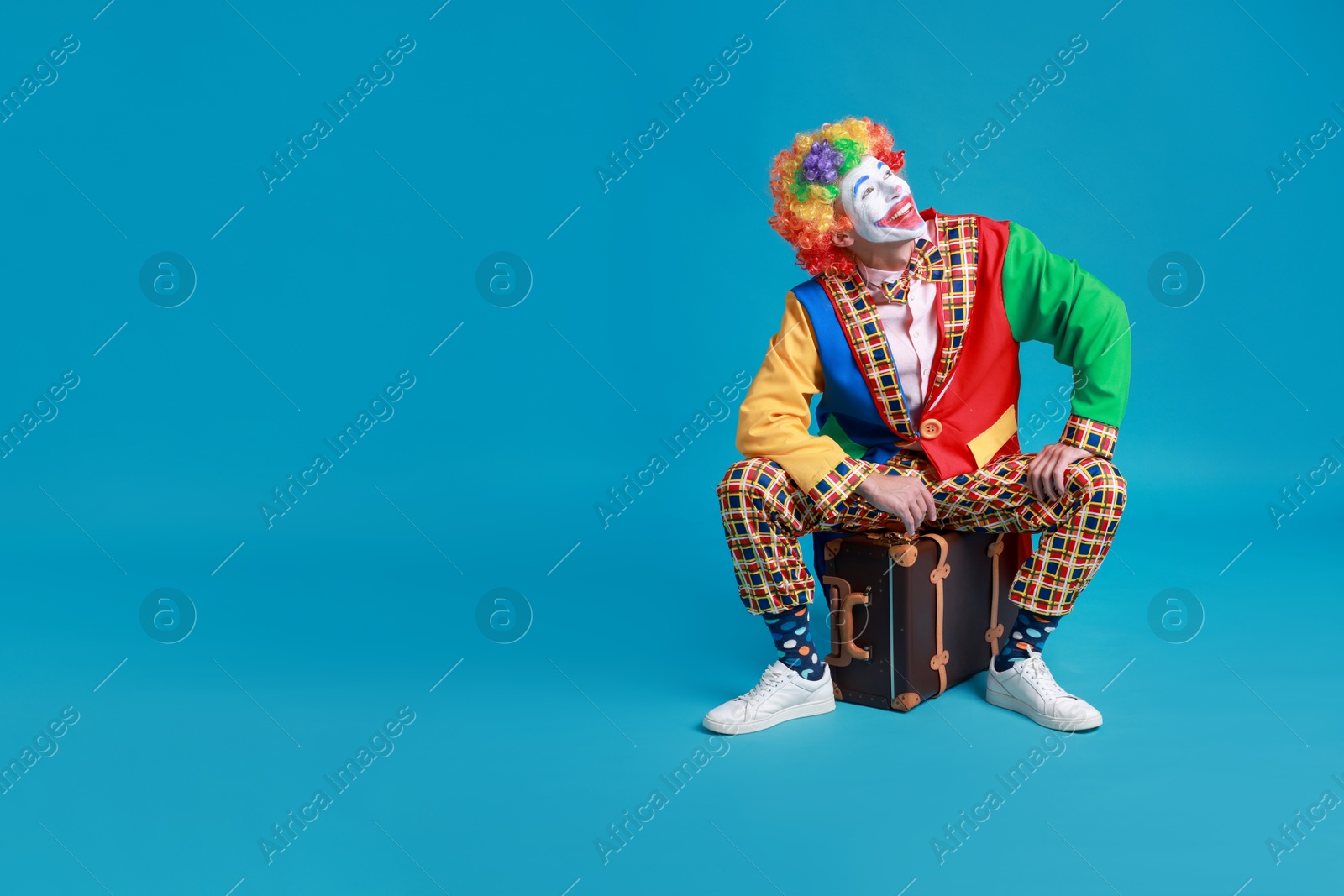 Photo of Happy clown sitting on suitcase against blue background, space for text