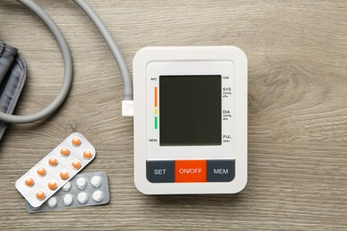 Photo of Blood pressure measuring device and pills on wooden table, flat lay