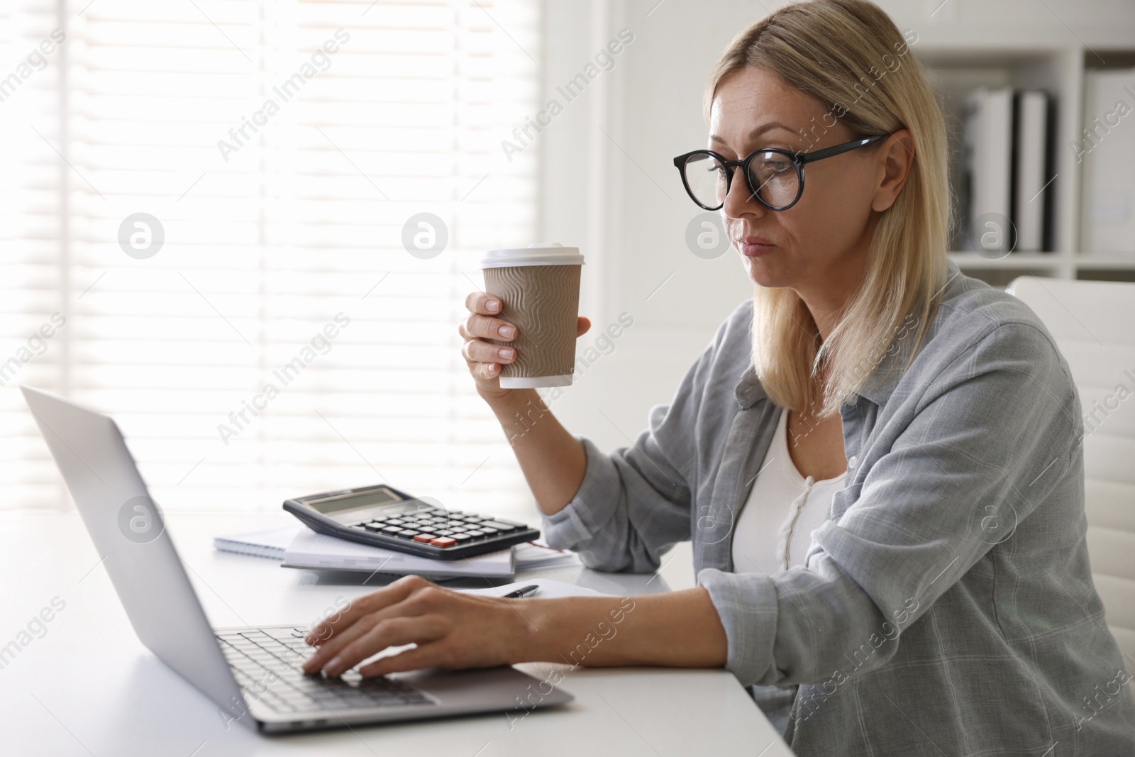 Photo of Budget planning. Woman with paper cup of coffee using laptop at table indoors