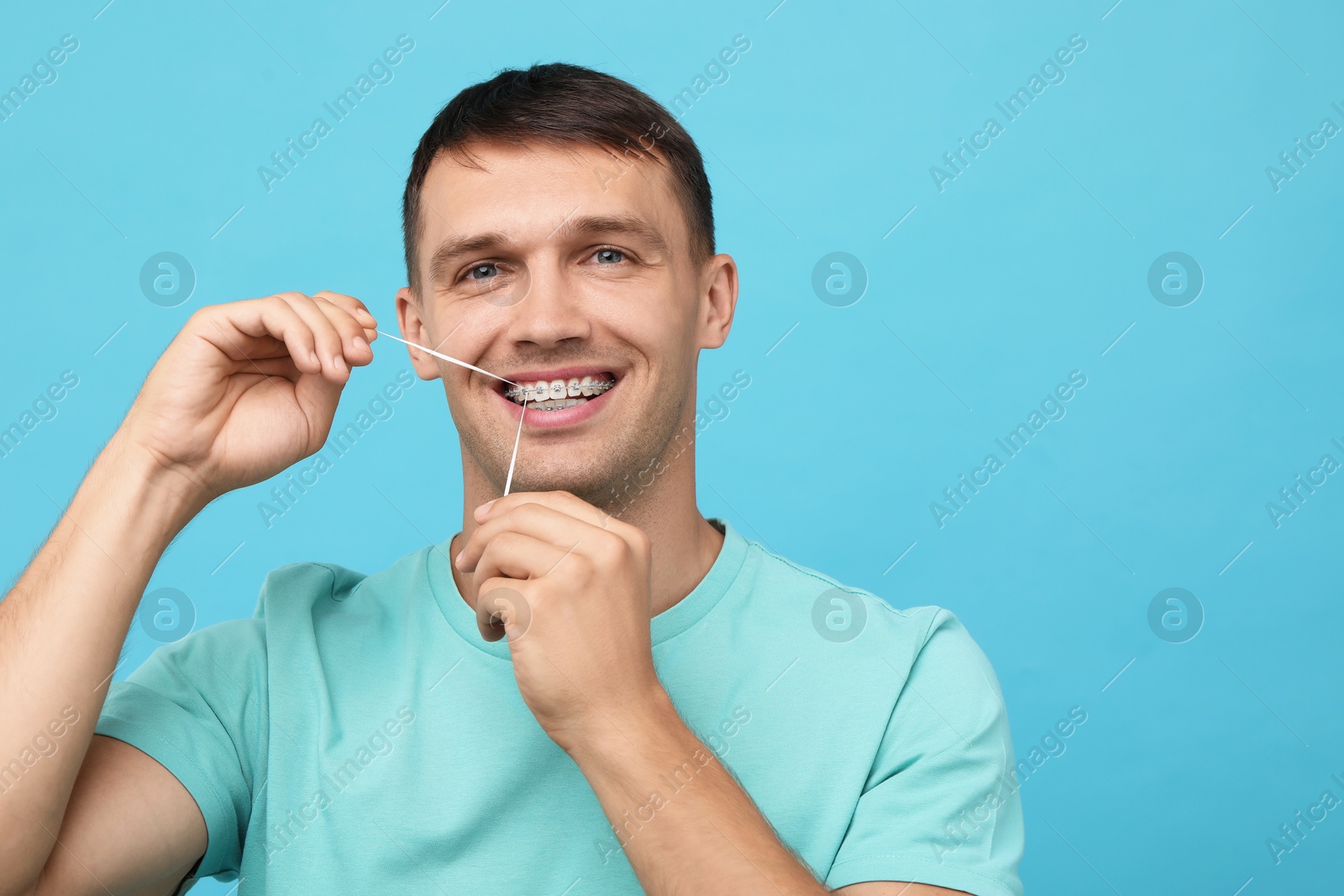 Photo of Man with braces cleaning teeth using dental floss on light blue background. Space for text