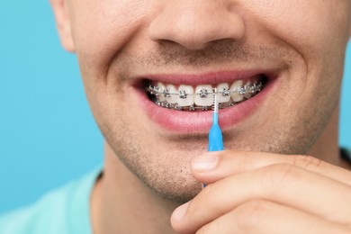 Photo of Man with dental braces cleaning teeth using interdental brush on light blue background, closeup