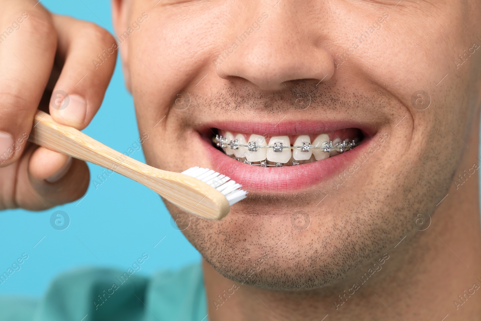 Photo of Man with dental braces cleaning teeth on light blue background, closeup