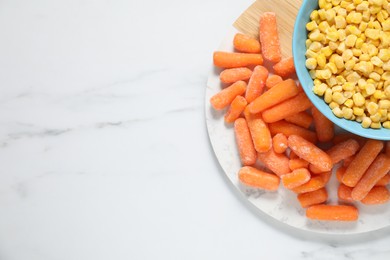 Photo of Frozen corn kernels in bowl and baby carrots on white marble table, top view. Space for text