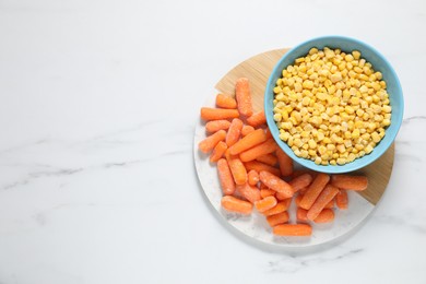 Photo of Frozen corn kernels in bowl and baby carrots on white marble table, top view. Space for text