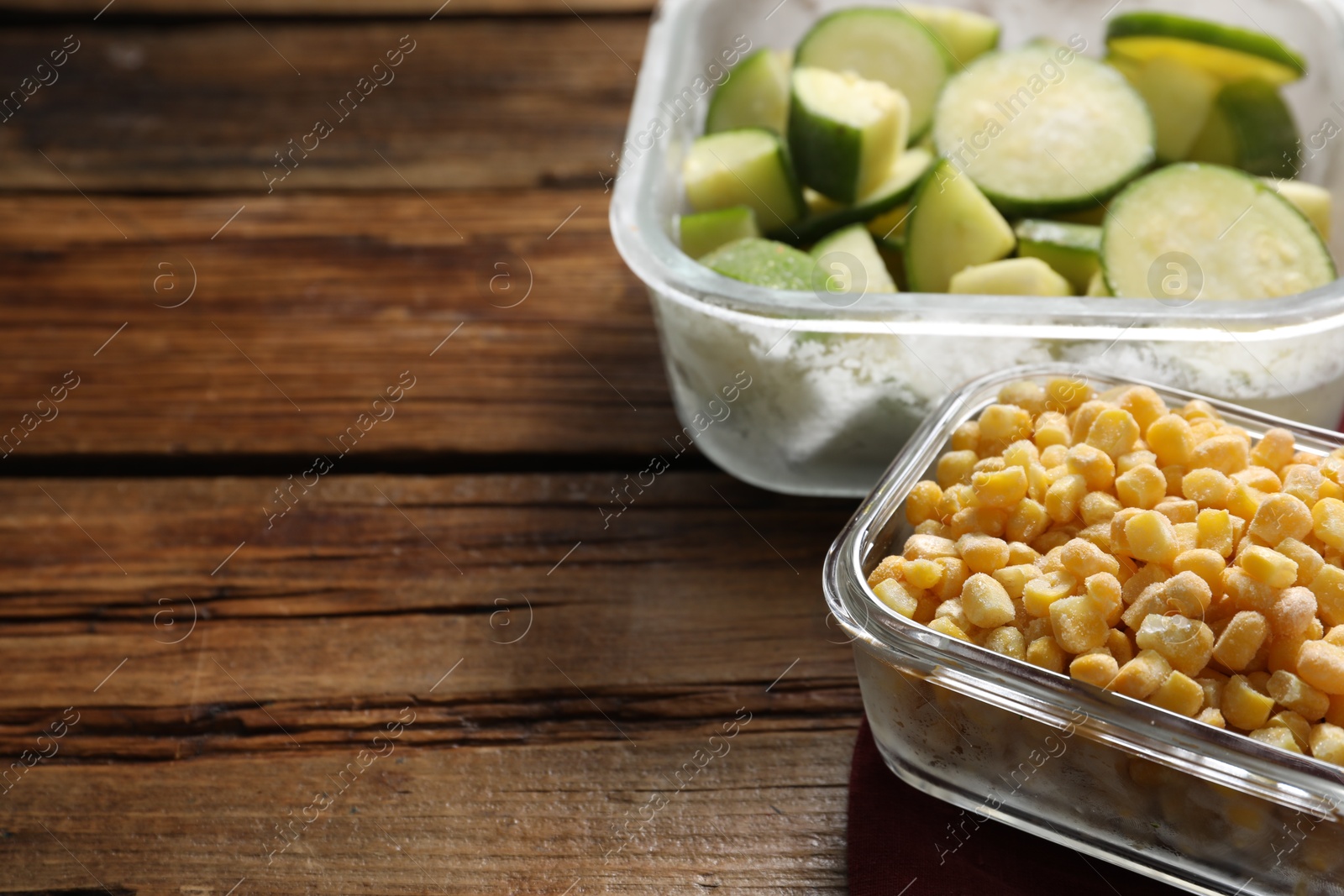 Photo of Frozen corn kernels and zucchini in glass containers on wooden table, closeup. Space for text