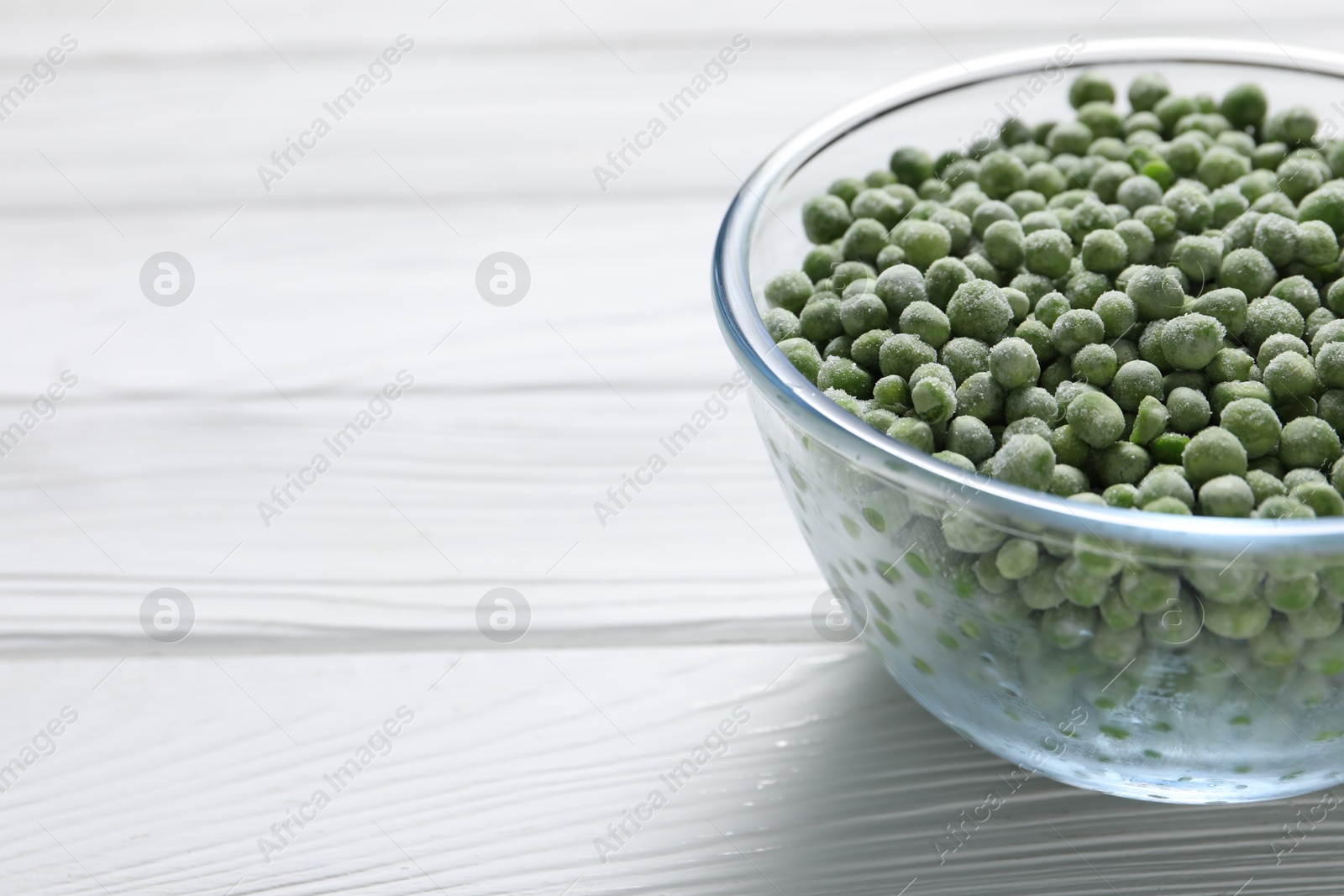 Photo of Frozen green peas in glass bowl on white wooden table, closeup. Space for text