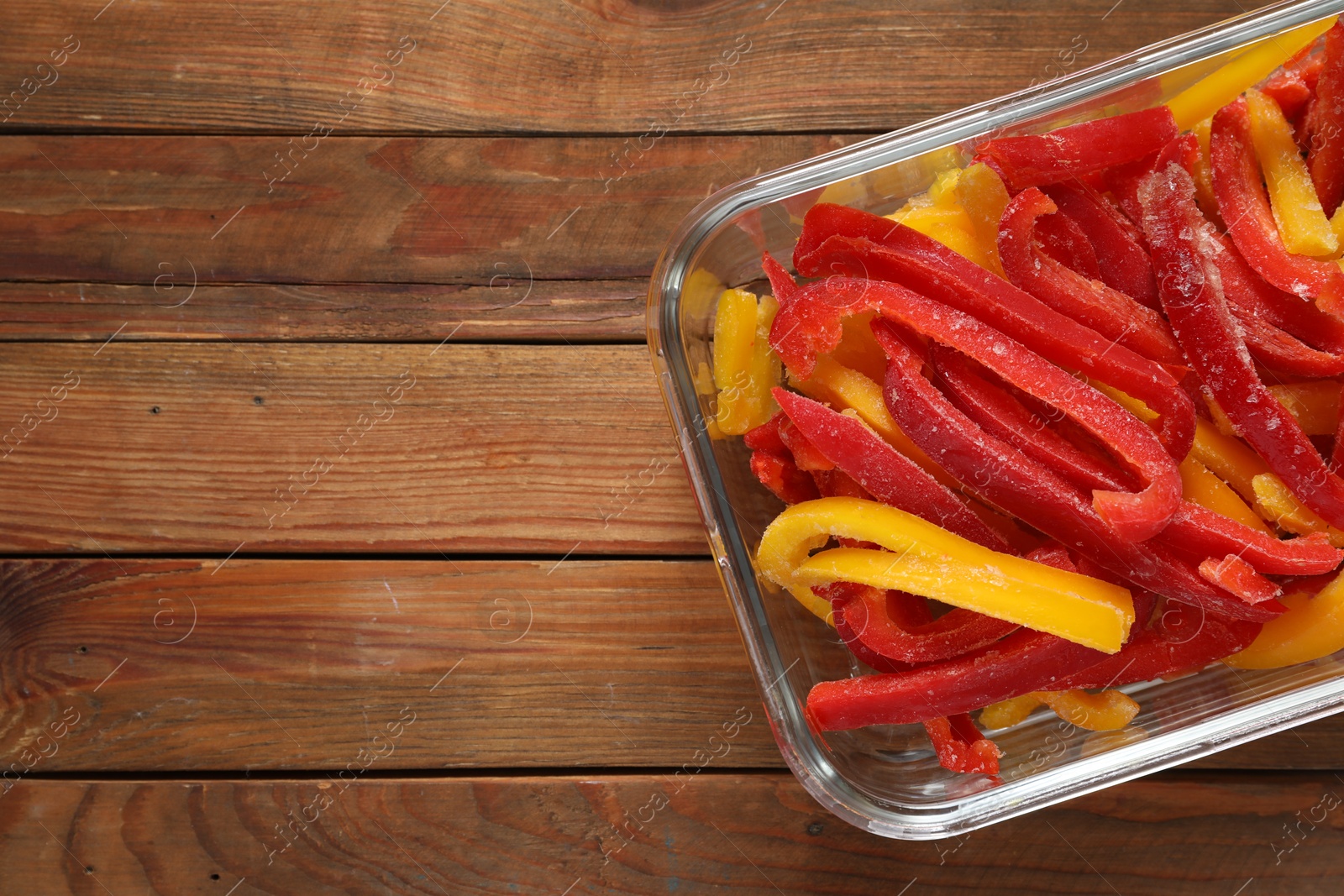 Photo of Slices of frozen bell peppers in glass container on wooden table, top view. Space for text