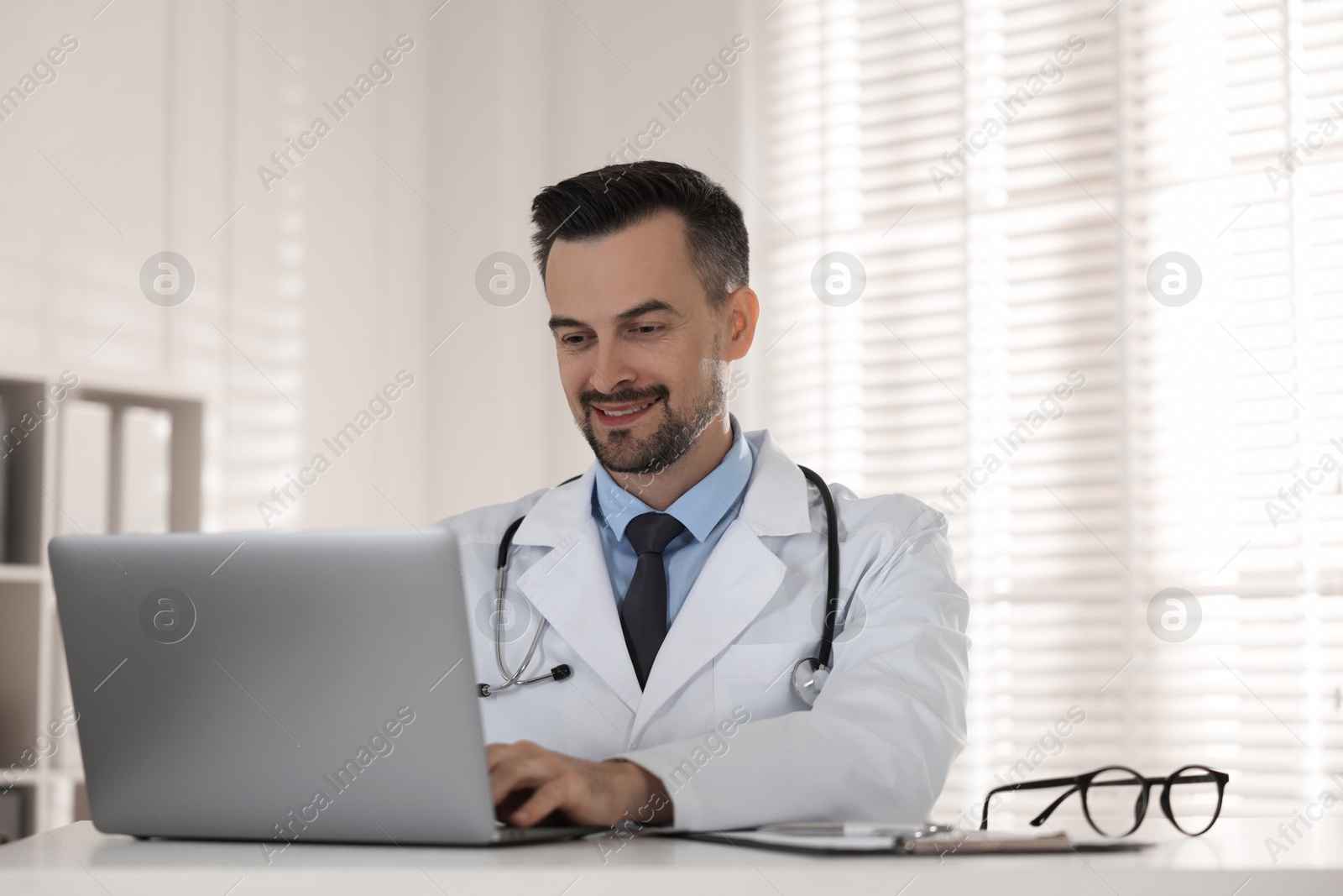 Photo of Smiling doctor working with laptop at table in clinic