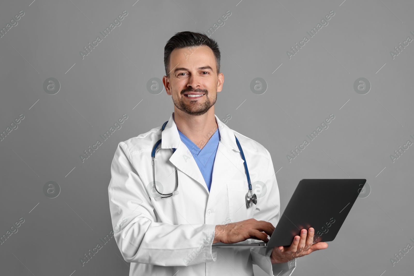 Photo of Smiling doctor with stethoscope and laptop on grey background