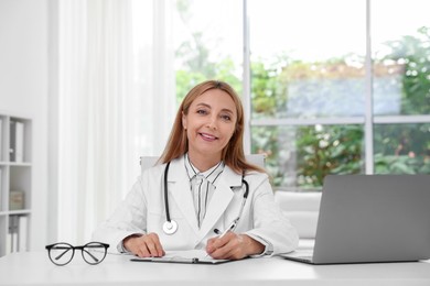 Doctor with stethoscope, laptop and clipboard at table in clinic