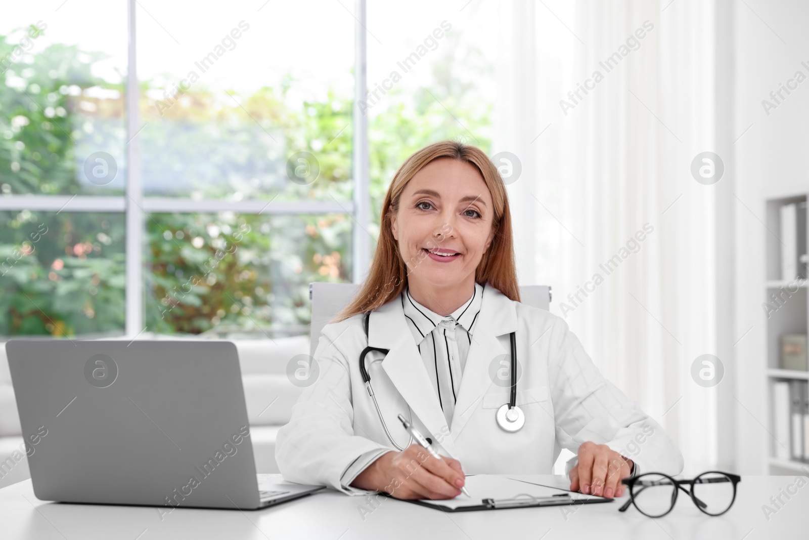 Photo of Doctor with stethoscope, laptop and clipboard at table in clinic