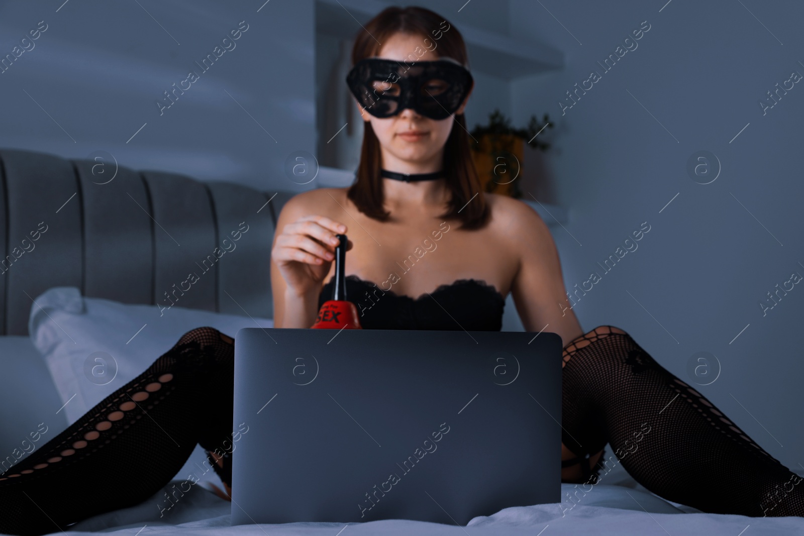 Photo of Prostitution. Webcam model in mask holding bell in front of laptop on bed indoors