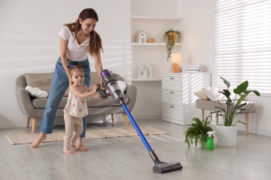 Photo of Smiling young woman and her daughter cleaning floor with cordless vacuum cleaner at home