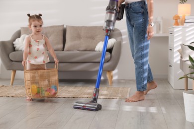 Photo of Young woman cleaning floor with cordless vacuum cleaner while her daughter playing with toys at home, closeup