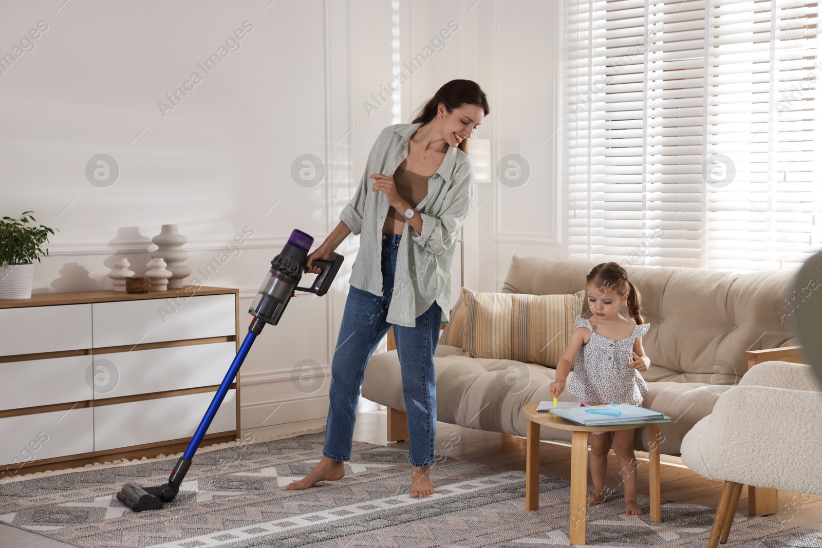 Photo of Smiling young woman cleaning rug with cordless vacuum cleaner while her daughter drawing at home