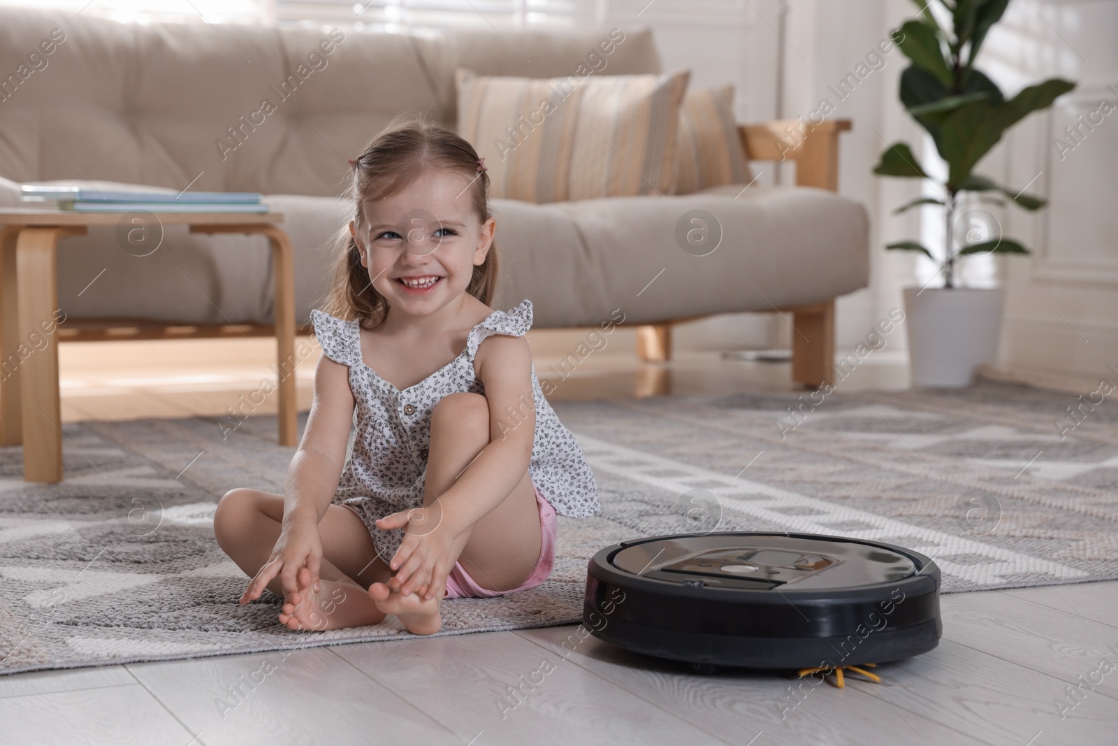 Photo of Cute little girl with robotic vacuum cleaner on floor at home