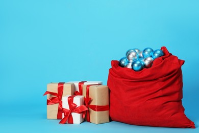 Photo of Santa Claus bag with baubles and gift boxes on light blue background, space for text