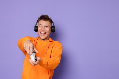 Photo of Happy young man in headphones playing video games with controller on violet background, space for text