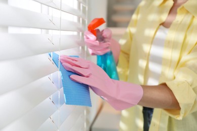 Young woman cleaning window blinds with rag and spray at home, closeup