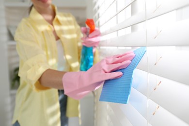 Young woman cleaning window blinds with rag and spray at home, closeup