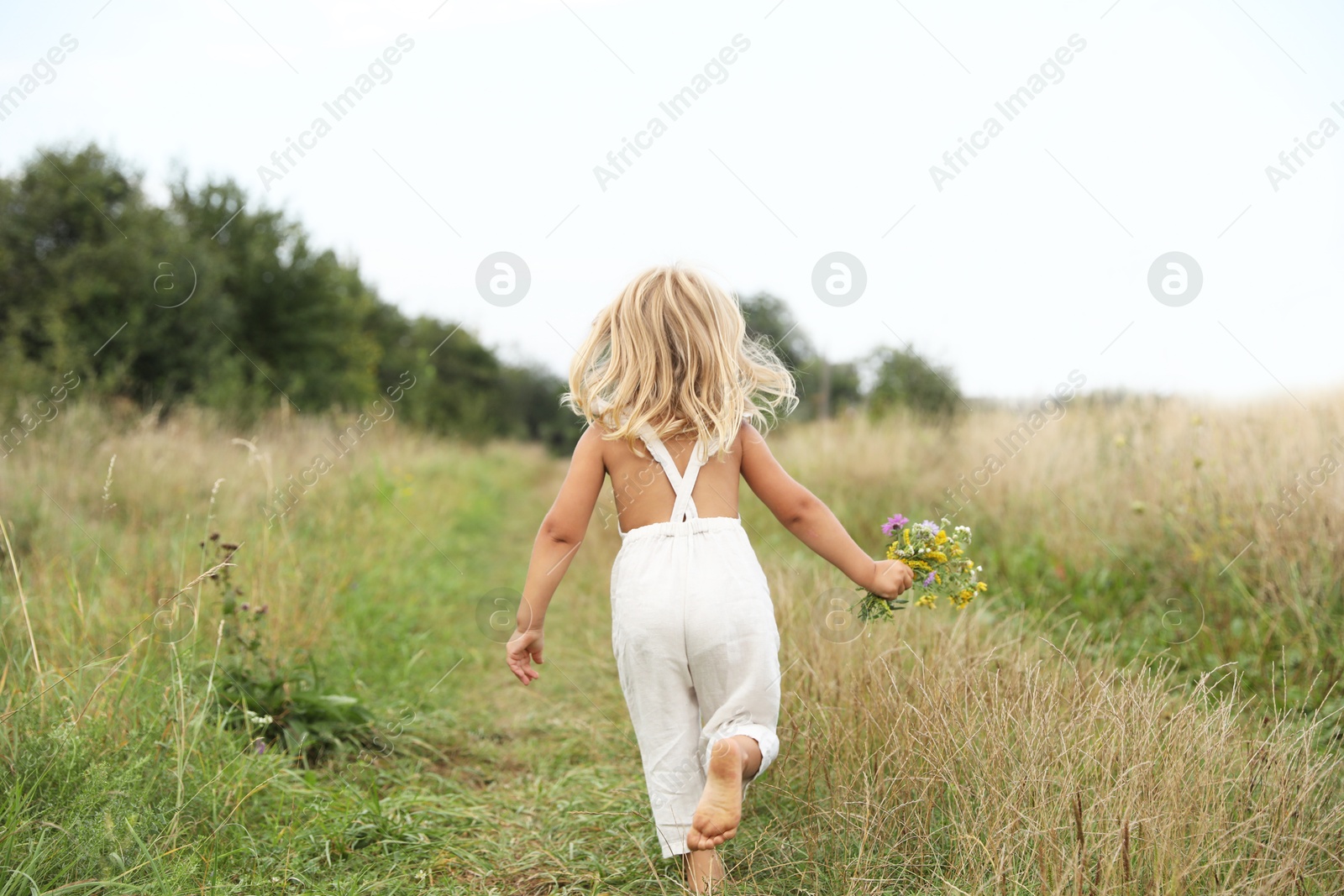 Photo of Little girl with flowers walking barefoot at meadow, back view. Child enjoying beautiful nature