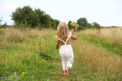 Photo of Little girl with flowers walking barefoot at meadow, back view. Child enjoying beautiful nature