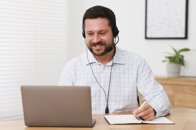 Interpreter in headset taking notes while having video chat via laptop at wooden table indoors