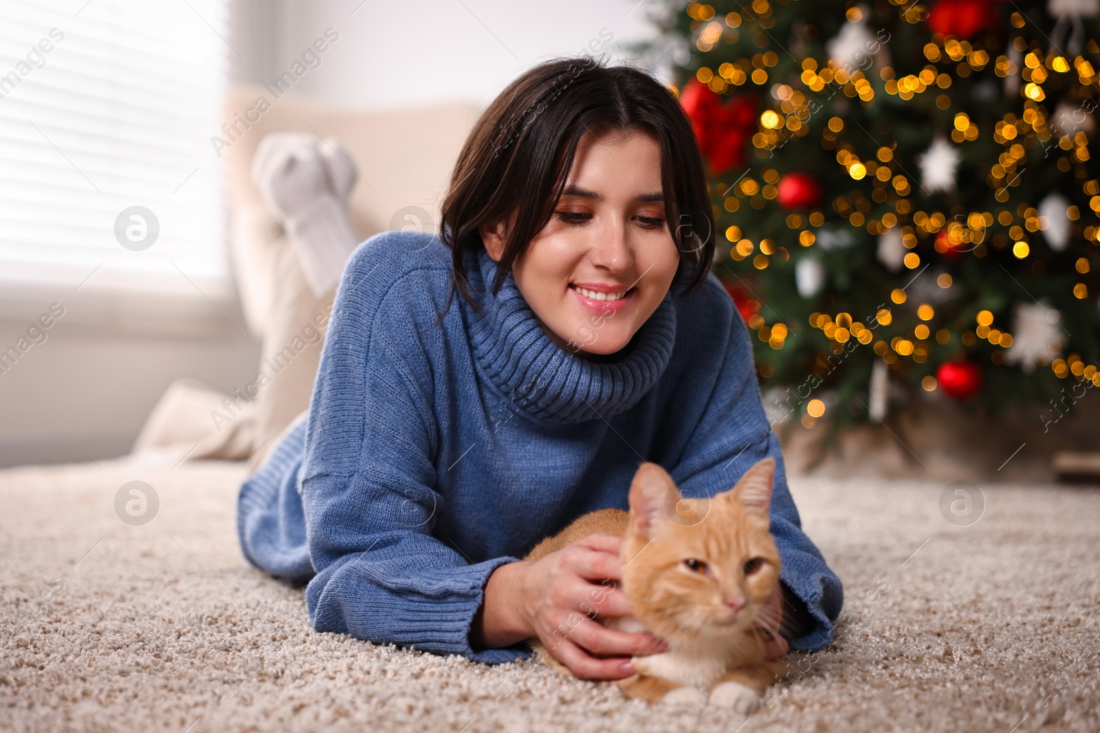 Photo of Woman with cute ginger cat lying on rug in room decorated for Christmas