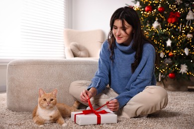 Photo of Woman opening Christmas gift and cute ginger cat lying on rug indoors