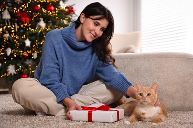 Photo of Woman with Christmas gift and cute ginger cat lying on rug indoors