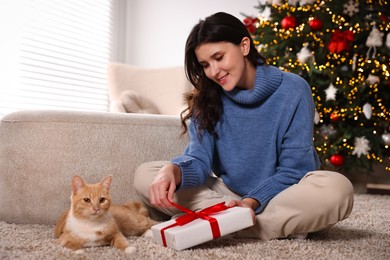 Woman with Christmas gift and cute ginger cat lying on rug indoors