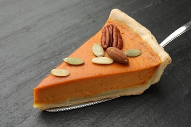 Photo of Piece of tasty homemade pumpkin pie with seeds, nuts and server on black table, closeup