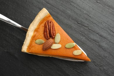 Piece of tasty homemade pumpkin pie with seeds, nuts and server on black table, top view