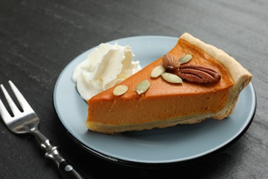 Photo of Piece of tasty homemade pumpkin pie with whipped cream, seeds, nuts and fork on black table, closeup