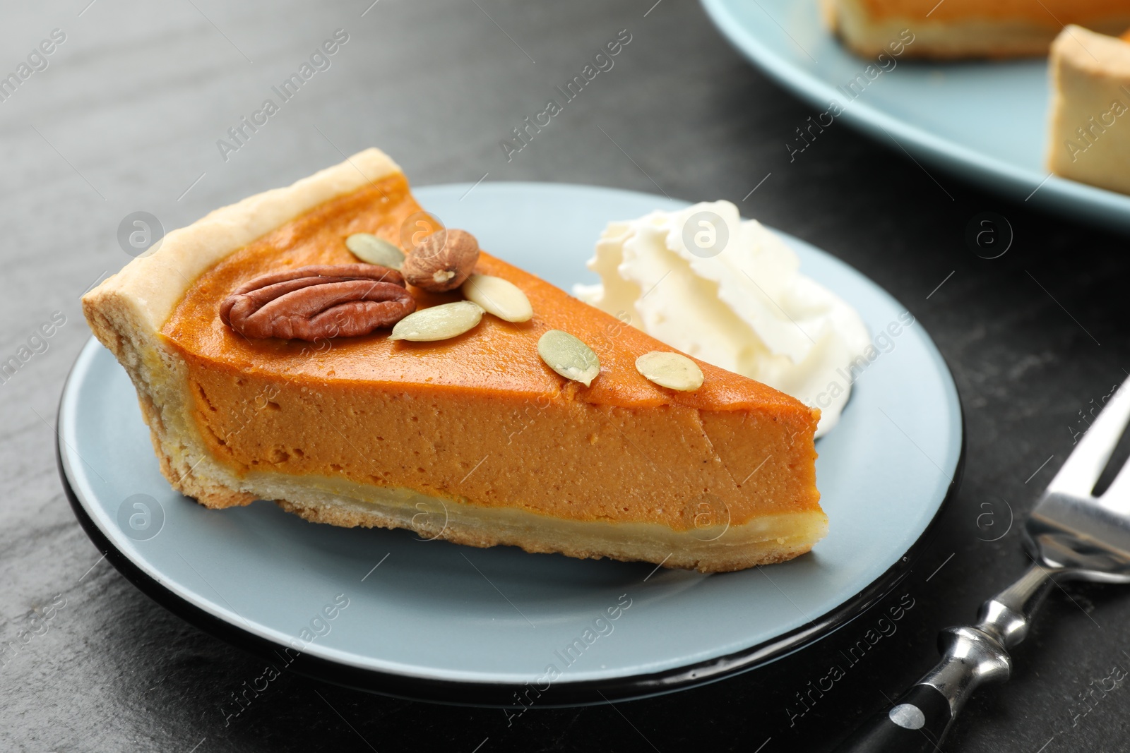 Photo of Piece of tasty homemade pumpkin pie with whipped cream, seeds, nuts and fork on black table, closeup