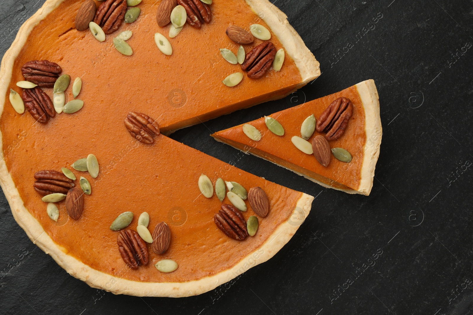 Photo of Tasty homemade pumpkin pie with seeds and nuts on black table, top view