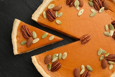 Photo of Tasty homemade pumpkin pie with seeds and nuts on black table, top view