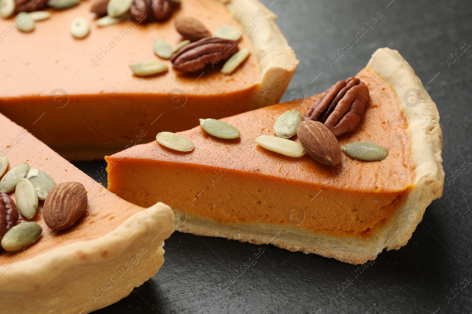 Photo of Pieces of tasty homemade pumpkin pie with seeds and nuts on black table, closeup