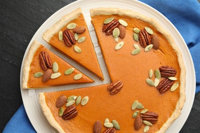 Photo of Tasty homemade pumpkin pie with seeds and nuts on black table, top view