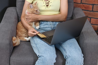 Online pet shop. Woman with laptop, credit card and cute cat making order on armchair at home, closeup