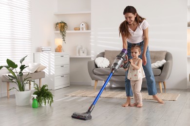 Photo of Smiling young woman and her daughter cleaning floor with cordless vacuum cleaner at home