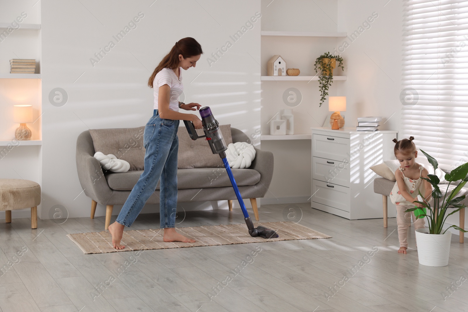 Photo of Young woman cleaning floor with cordless vacuum cleaner while her daughter spraying houseplant at home