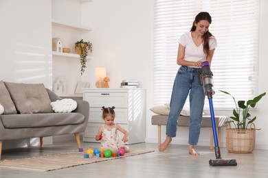 Smiling young woman cleaning floor with cordless vacuum cleaner while her daughter playing with toys at home