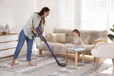 Photo of Smiling young woman cleaning rug with cordless vacuum cleaner while her daughter drawing at home