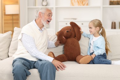 Grandpa and his granddaughter playing with toy bear on sofa at home