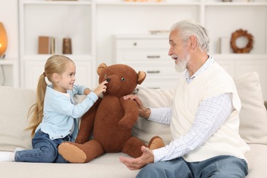 Grandpa and his granddaughter playing with toy bear on sofa at home