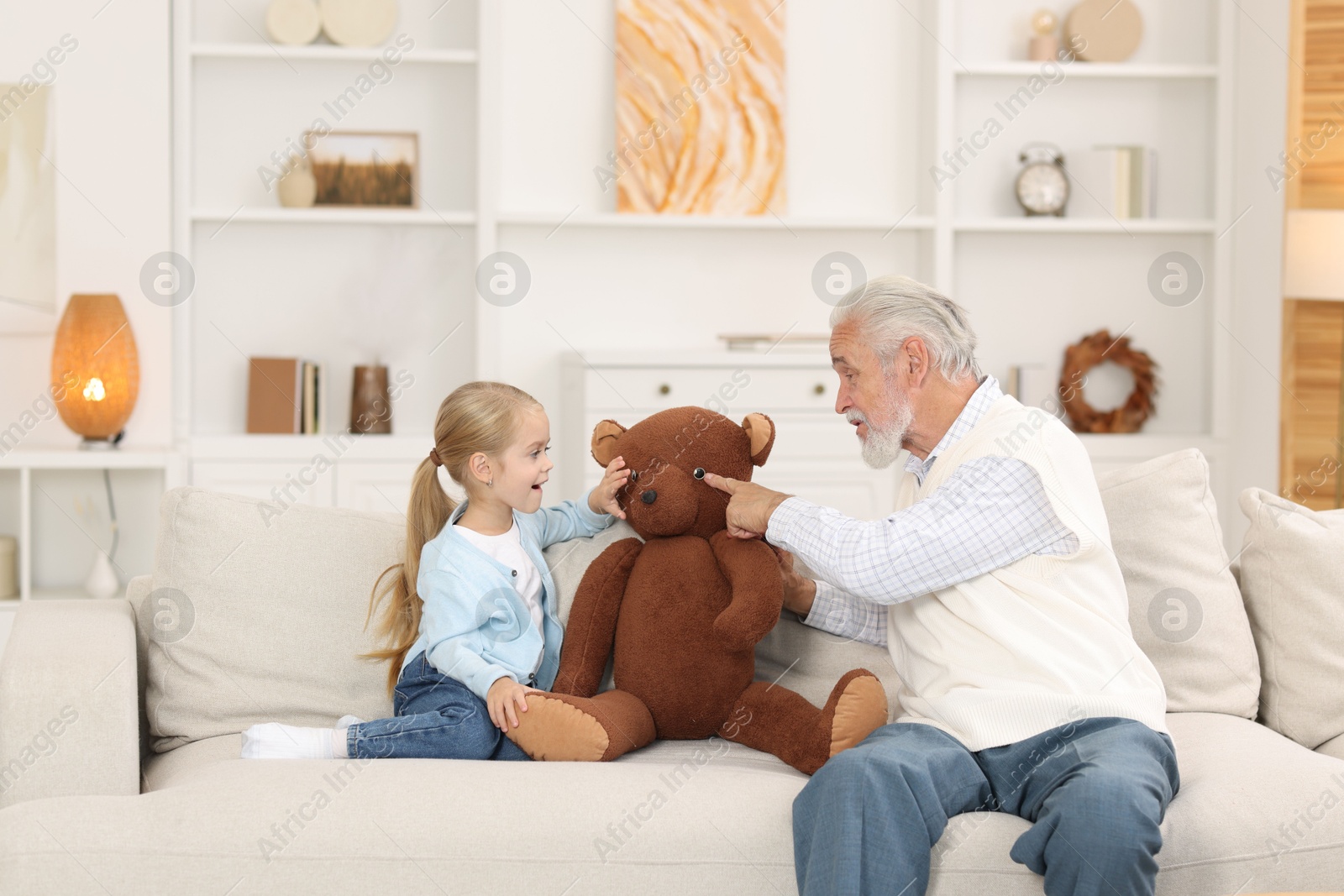 Photo of Grandpa and his granddaughter playing with toy bear on sofa at home