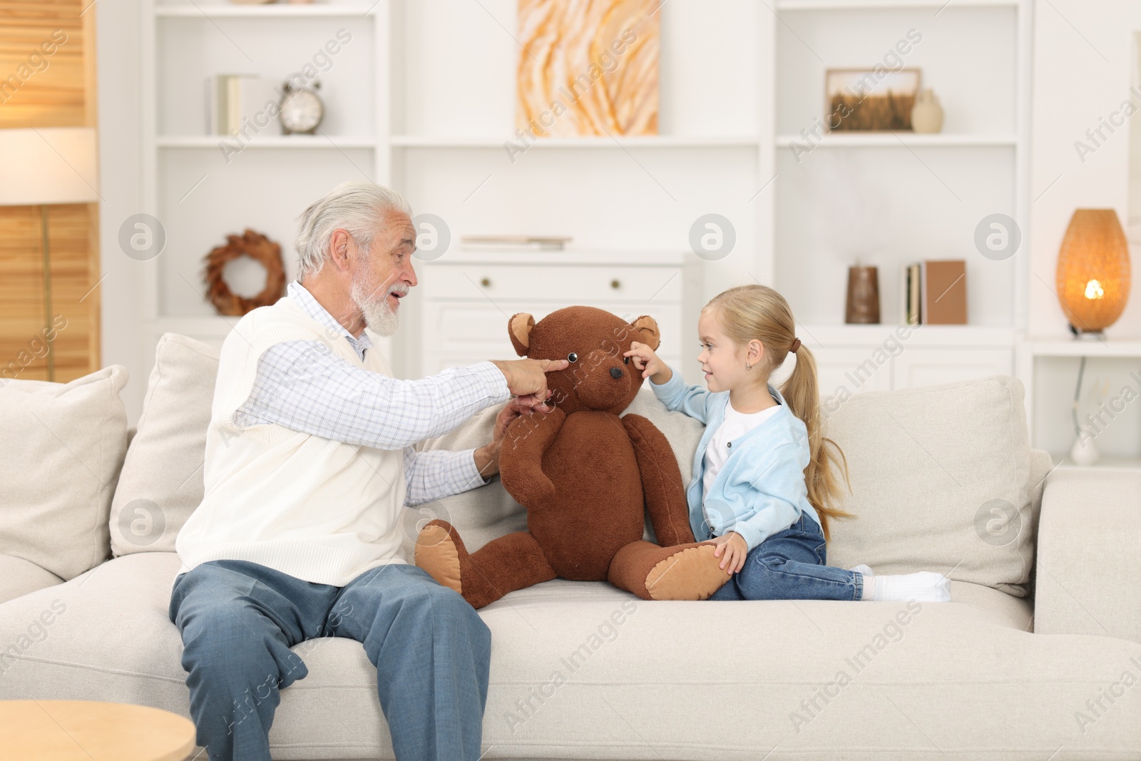 Photo of Grandpa and his granddaughter playing with toy bear on sofa at home