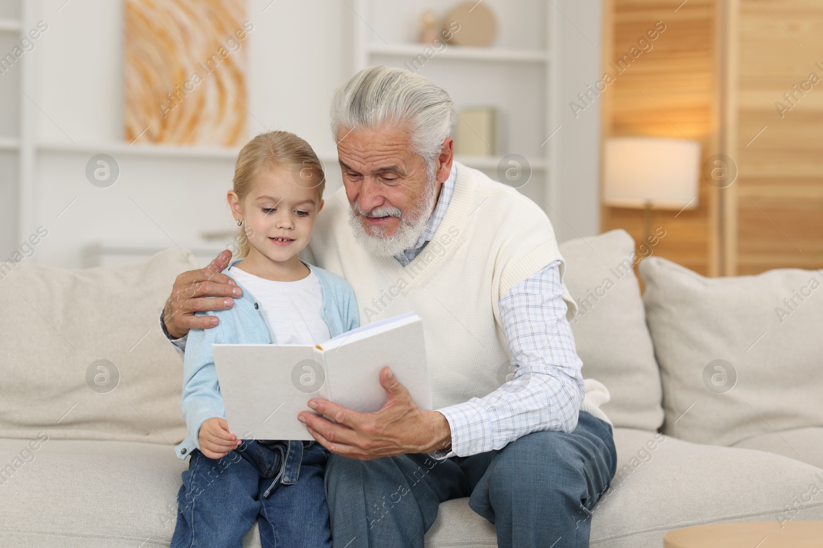 Photo of Grandpa and his granddaughter reading book together on sofa at home